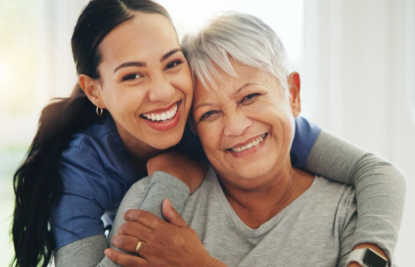 24-hour caregiver warmly cuddles elderly lady, both smiling and enjoying a moment together.